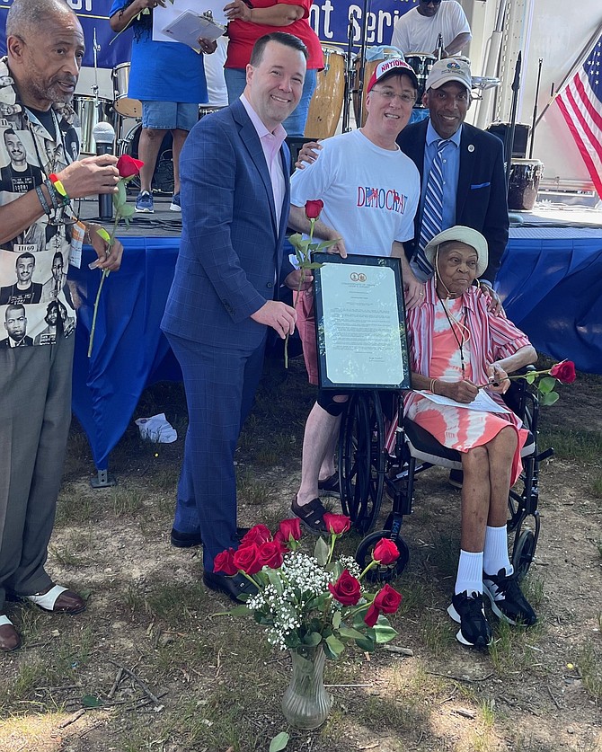 New Gum Springs Civic Association President Vince Carter (left) praises Mattie Palmore (seated) before adding a rose to her bouquet. Mount Vernon School Board Member Mateo Dunn, Del. Paul Krizek (holding the General Assembly resolution), and Franconia Supervisor Rodney Lusk stand behind her.